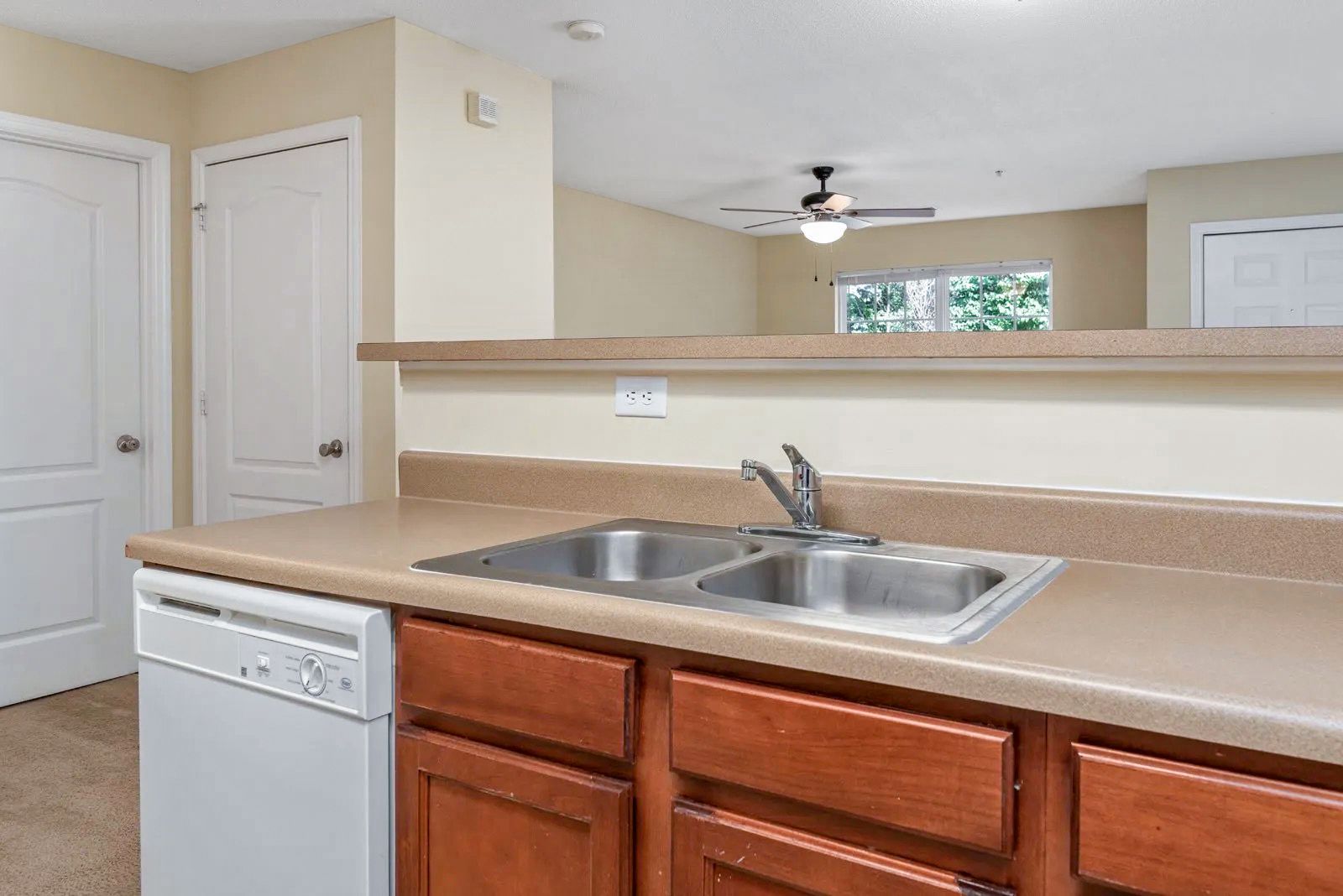 Kitchen with brown cabinets, a double sink, and a white dishwasher.  A doorway leads to a living area.