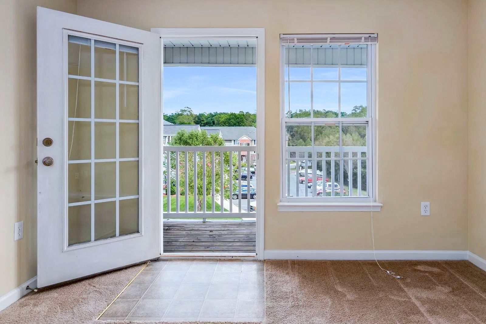 Open white French door and window revealing balcony and outside view; beige walls, brown carpet.