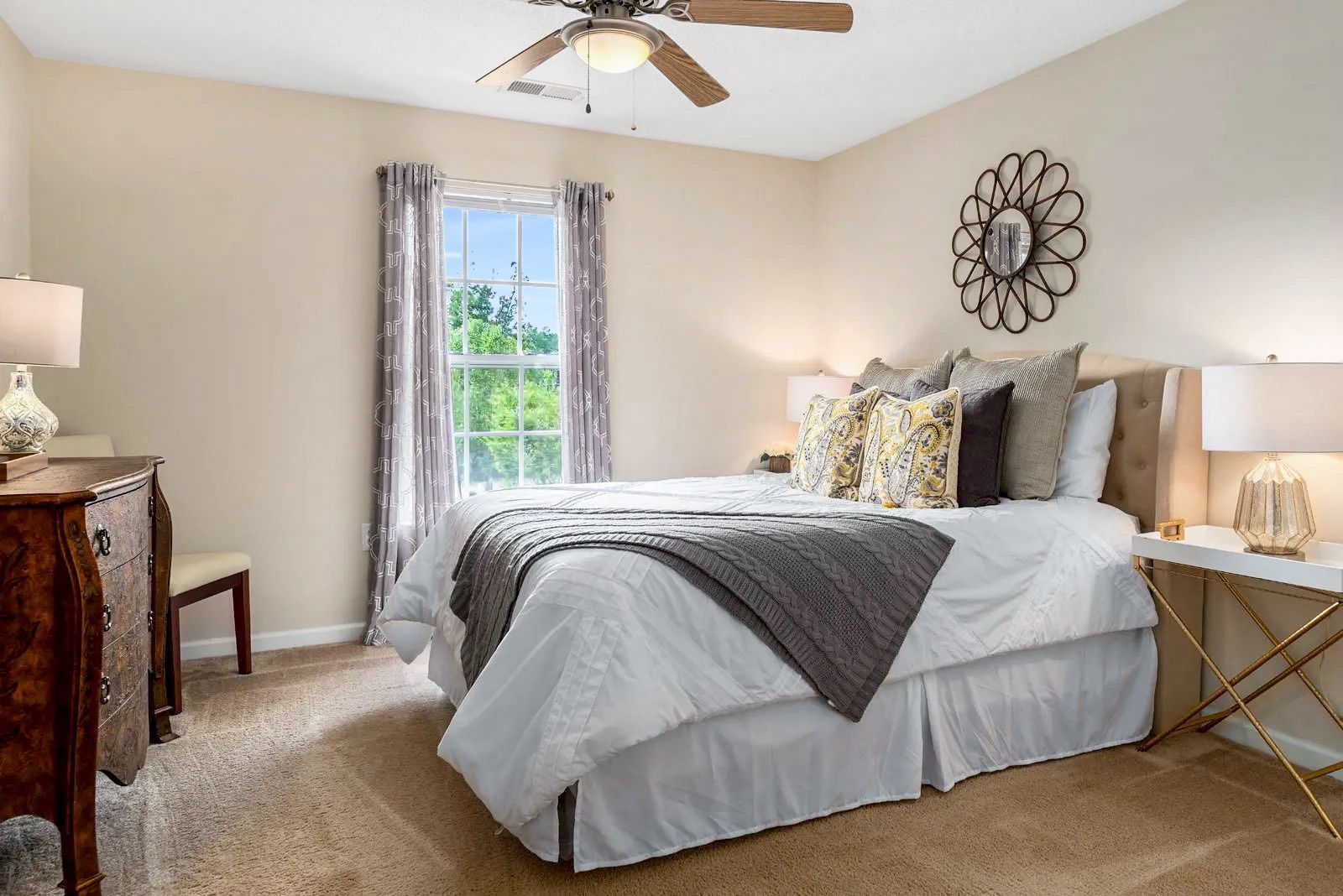 Bedroom with bed, dresser, window, and decorative mirror. Beige walls and carpet. White and gold accents.