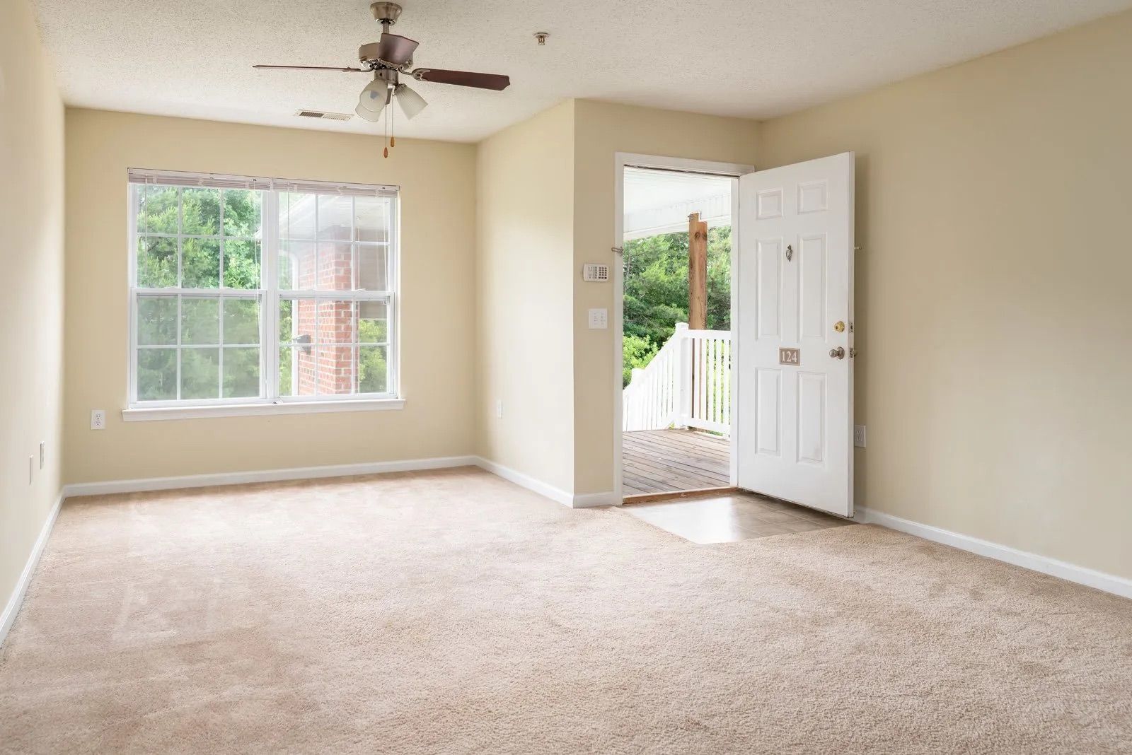 Empty living room with tan carpet, a window, and an open doorway to a porch. Beige walls and a ceiling fan.