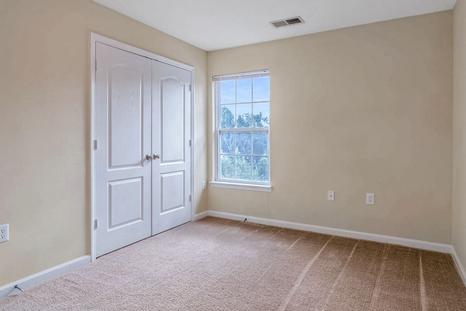 Empty bedroom with beige walls, a closed white closet, window with natural light, and carpet flooring.