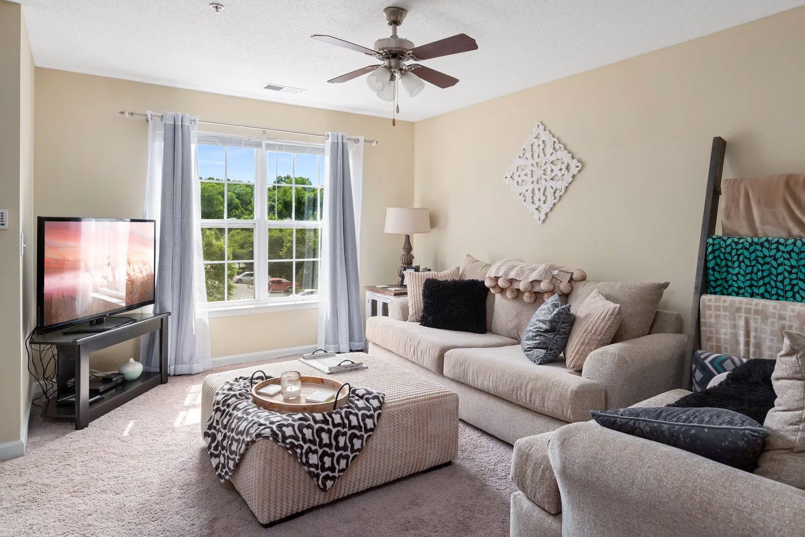Living room with beige sofa, patterned ottoman, TV, and window with curtains.