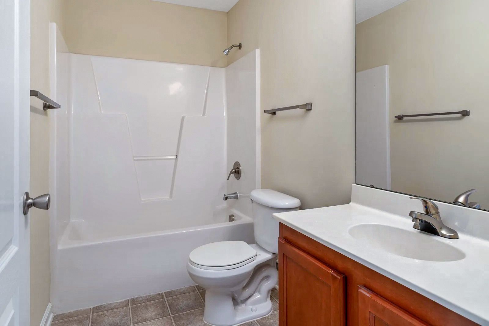 Bathroom with a white bathtub, toilet, and sink with wooden cabinet. Beige walls, silver towel racks.