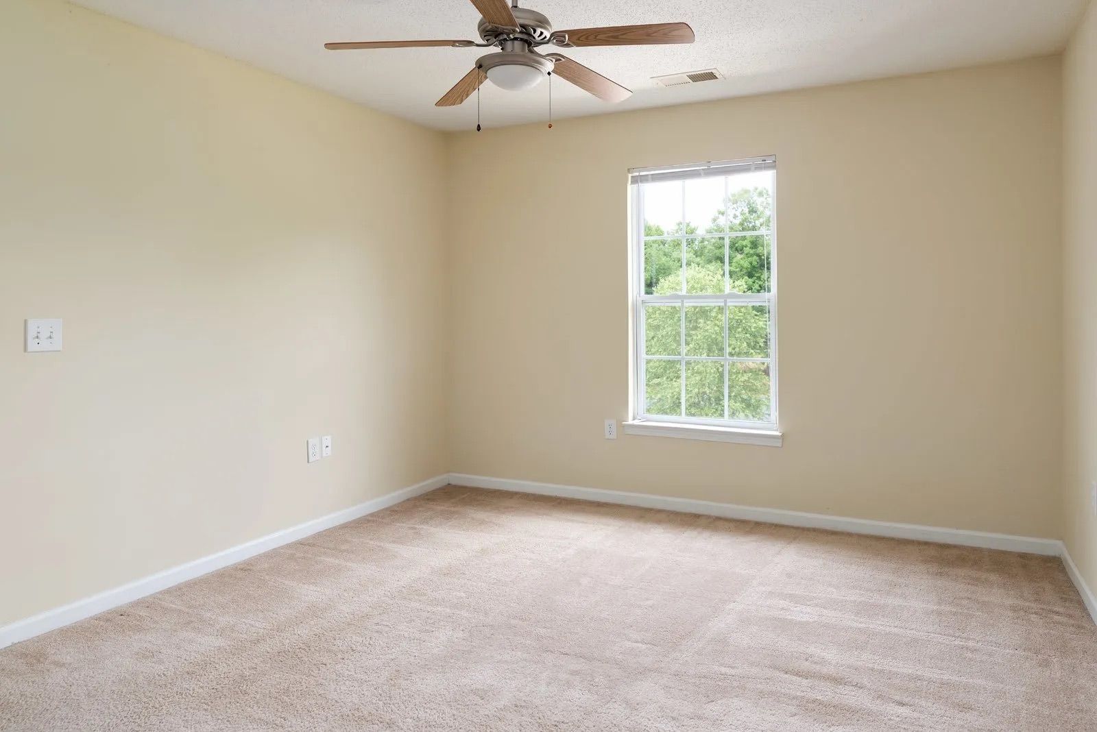 Empty beige bedroom with a window, ceiling fan, and carpeted floor.