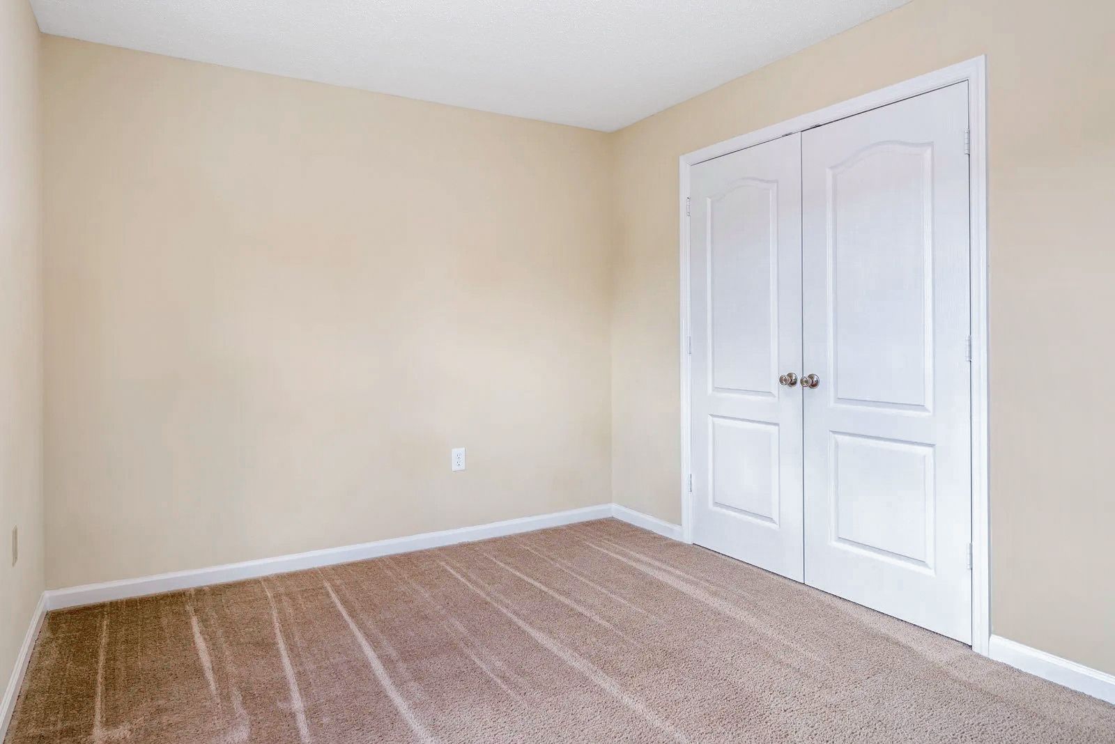 Empty room with beige walls, white double doors, and brown carpet.
