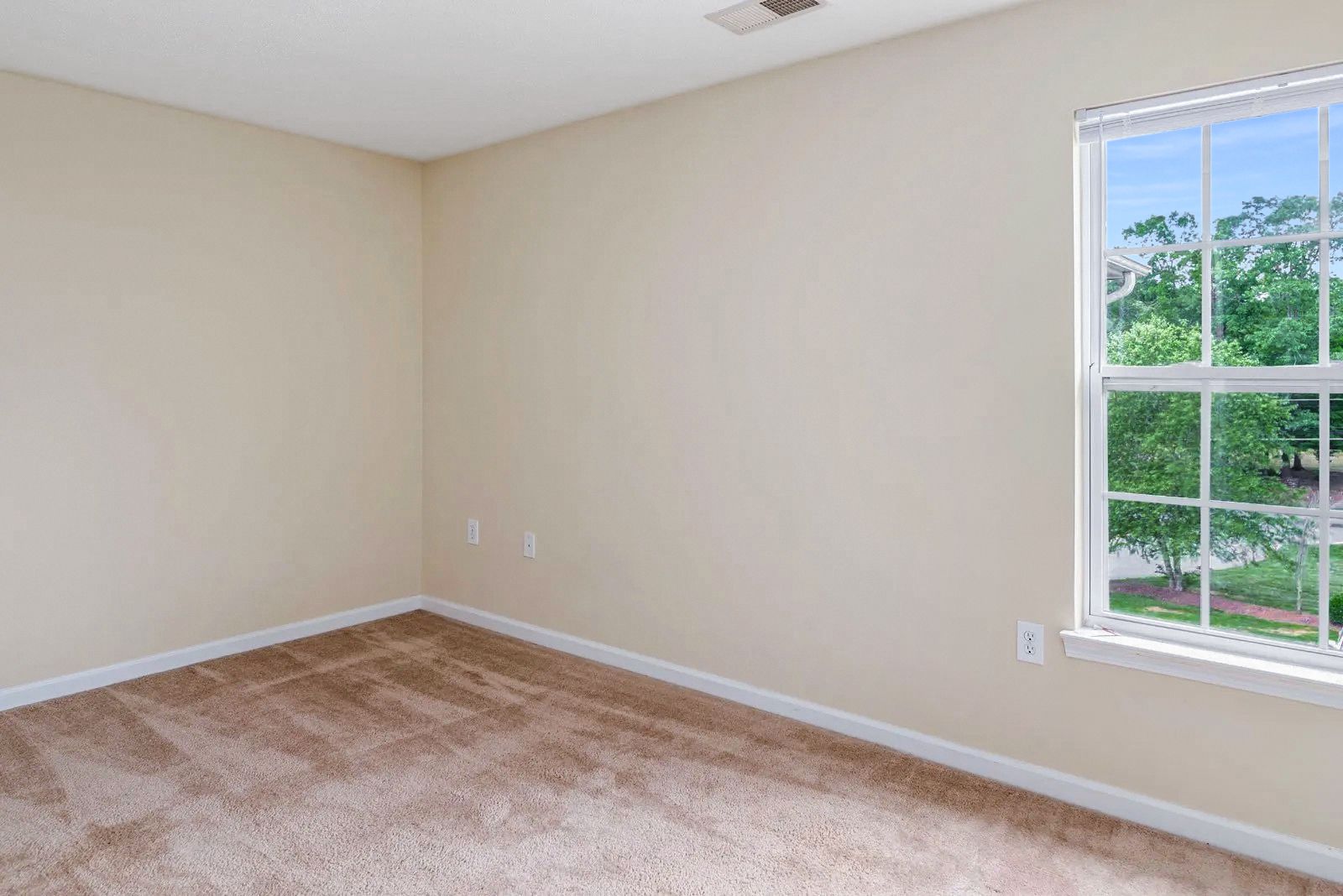 Empty bedroom with tan walls, brown carpet, and a window with a view of trees.