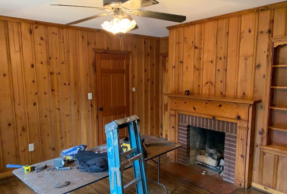 A living room with wood paneling , a fireplace and a ceiling fan.