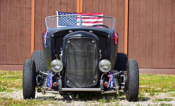 A black hot rod car parked on gravel in front of a wood-paneled building with an American flag displayed on the windshield.