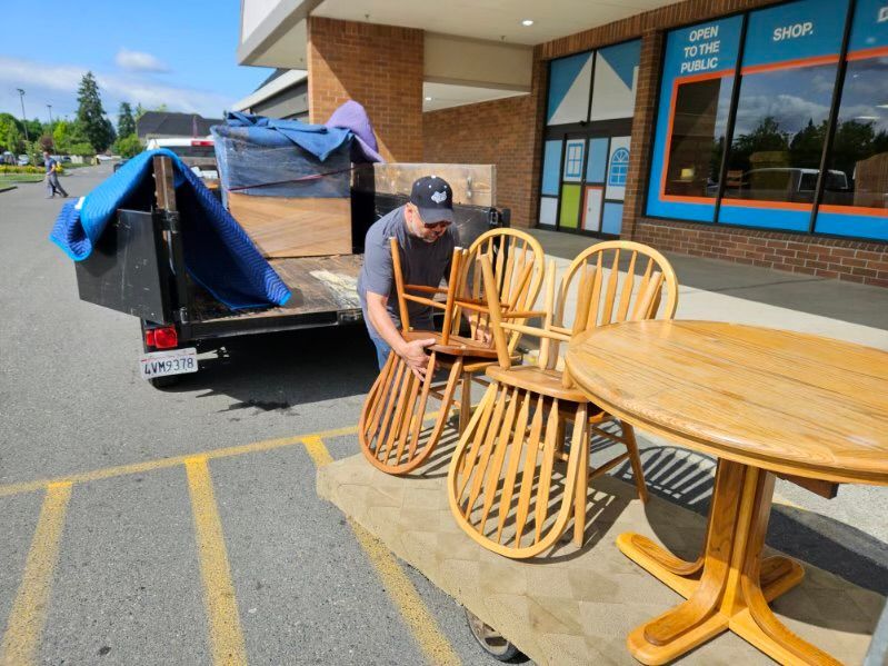 Man loading wooden chairs and table into a trailer outside a store.