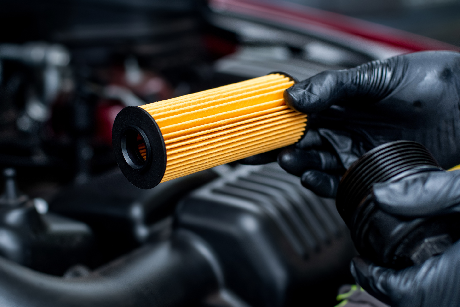 A person wearing black gloves is holding an oil filter in front of a car engine.