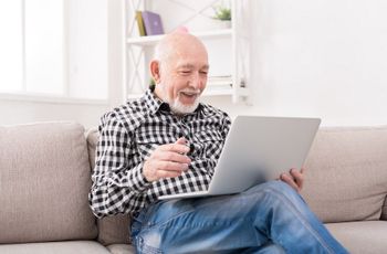 Older gentleman sitting on a sofa with an open laptop on his lap and a pair of glasses in his hand representing IT support required by home users