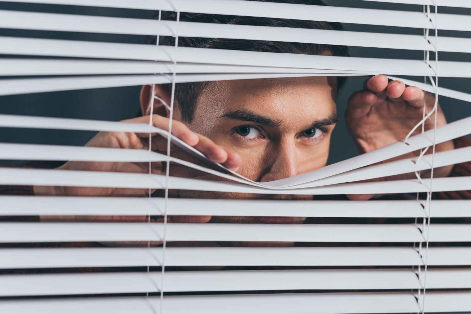 A man pulling apart some blinds to peer through suspiciously