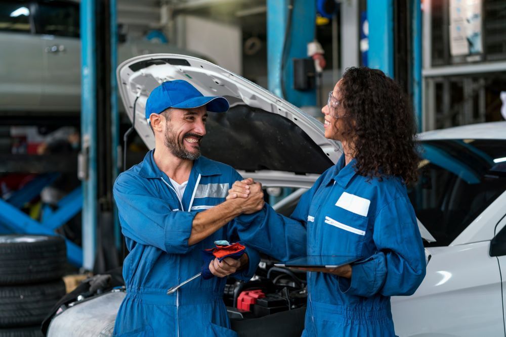 Two automotive mechanics smiling and clapping hands in front of a car with its bonnet open