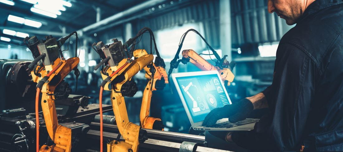 Man working on a laptop controlling manufacturing robotic arms
