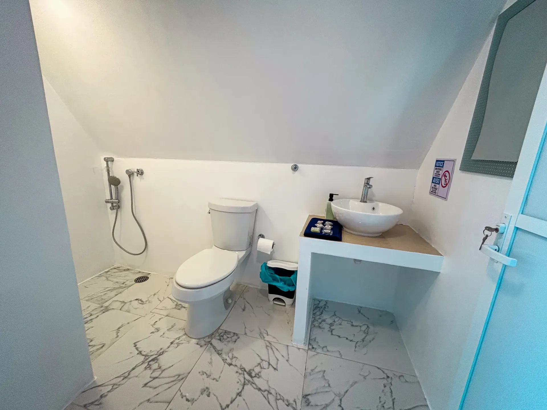 Baño con inodoro, lavabo y ducha bajo techo inclinado. Paredes blancas y suelo con estampado de mármol.