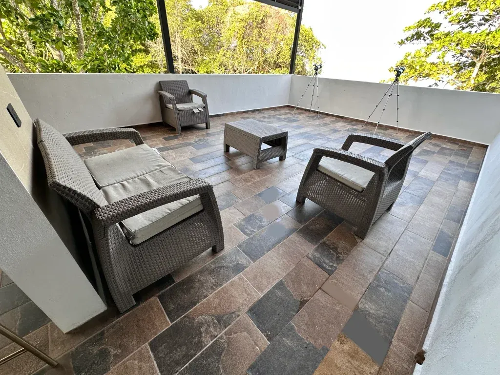 Patio con muebles de mimbre gris, suelo de baldosas marrones y vistas a los árboles.