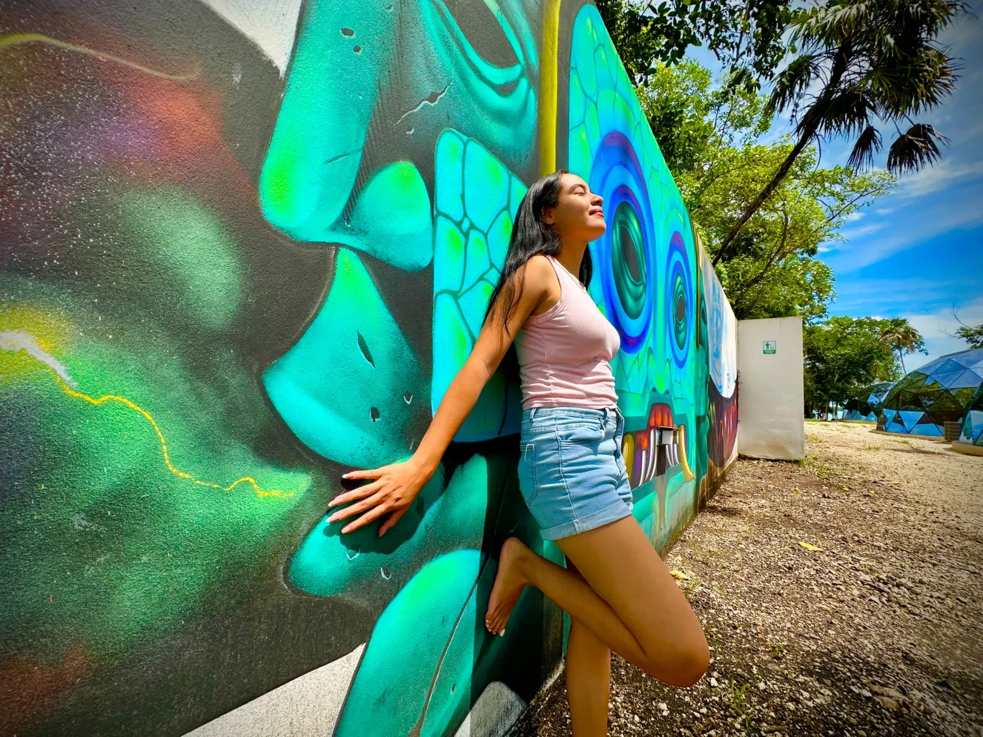 Una mujer en pantalones cortos y camiseta de tirantes se apoya en un colorido mural al aire libre en un día soleado.