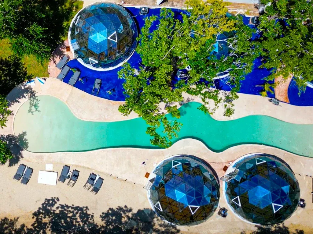 Vista aérea de un complejo turístico con cúpulas geodésicas, piscina y tumbonas rodeadas de vegetación.