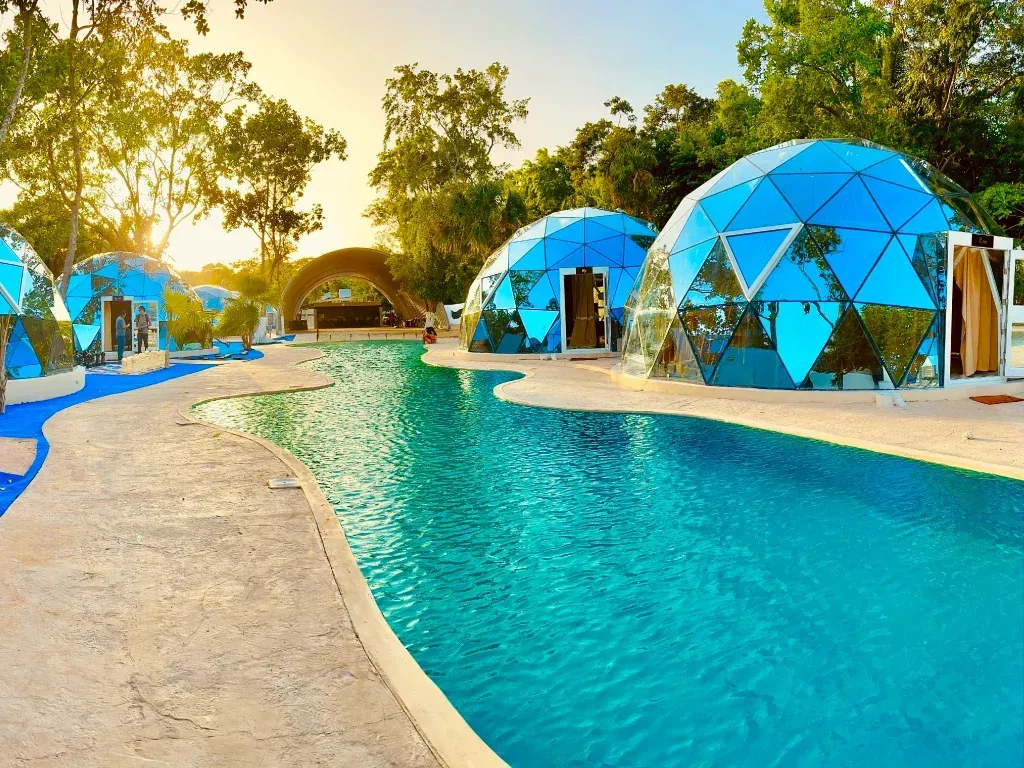 Estructuras de cúpulas geodésicas azules junto a una piscina turquesa, en un entorno soleado al aire libre.
