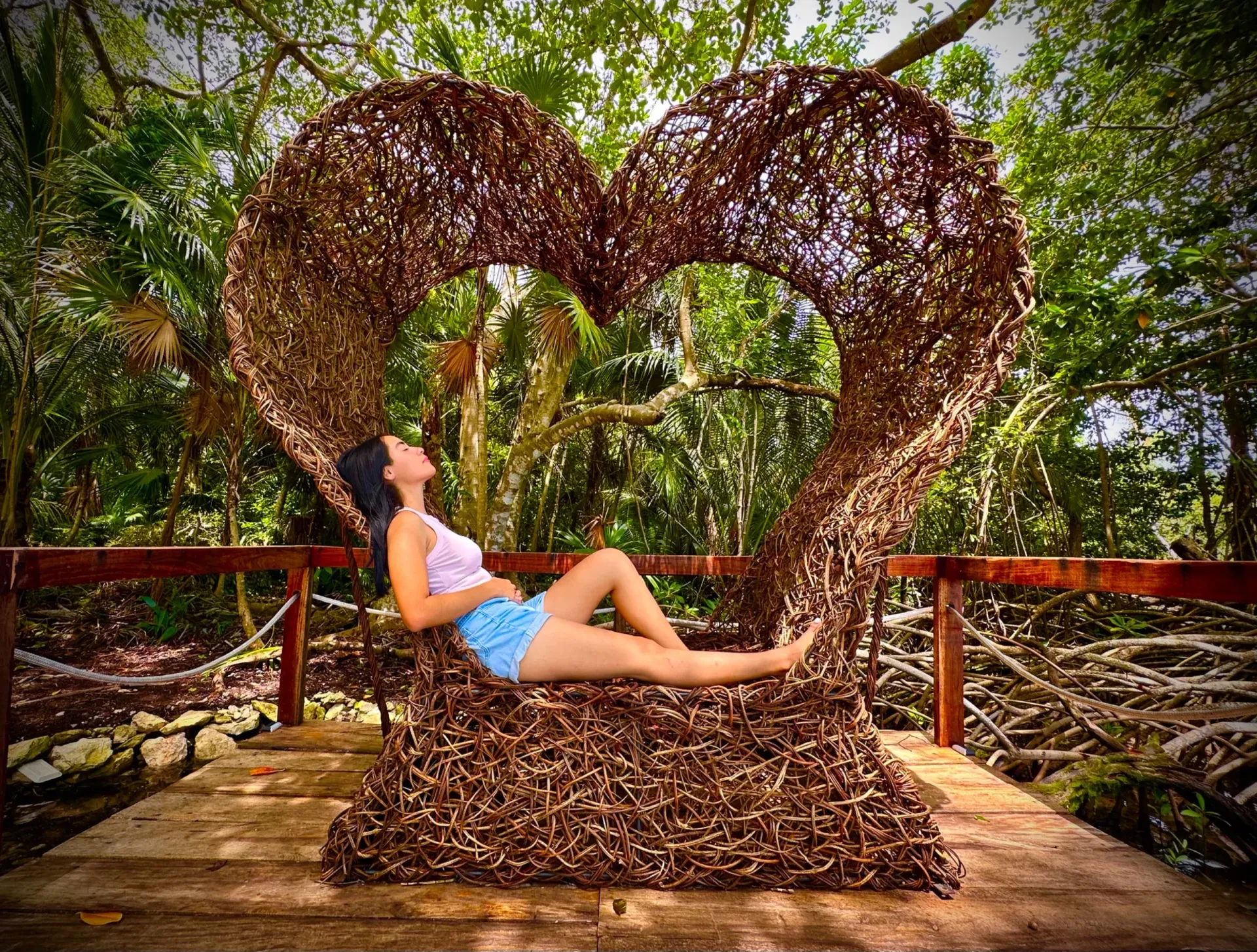 Una mujer yace reclinada en una gran estructura tejida en forma de corazón sobre una plataforma de madera rodeada de árboles.