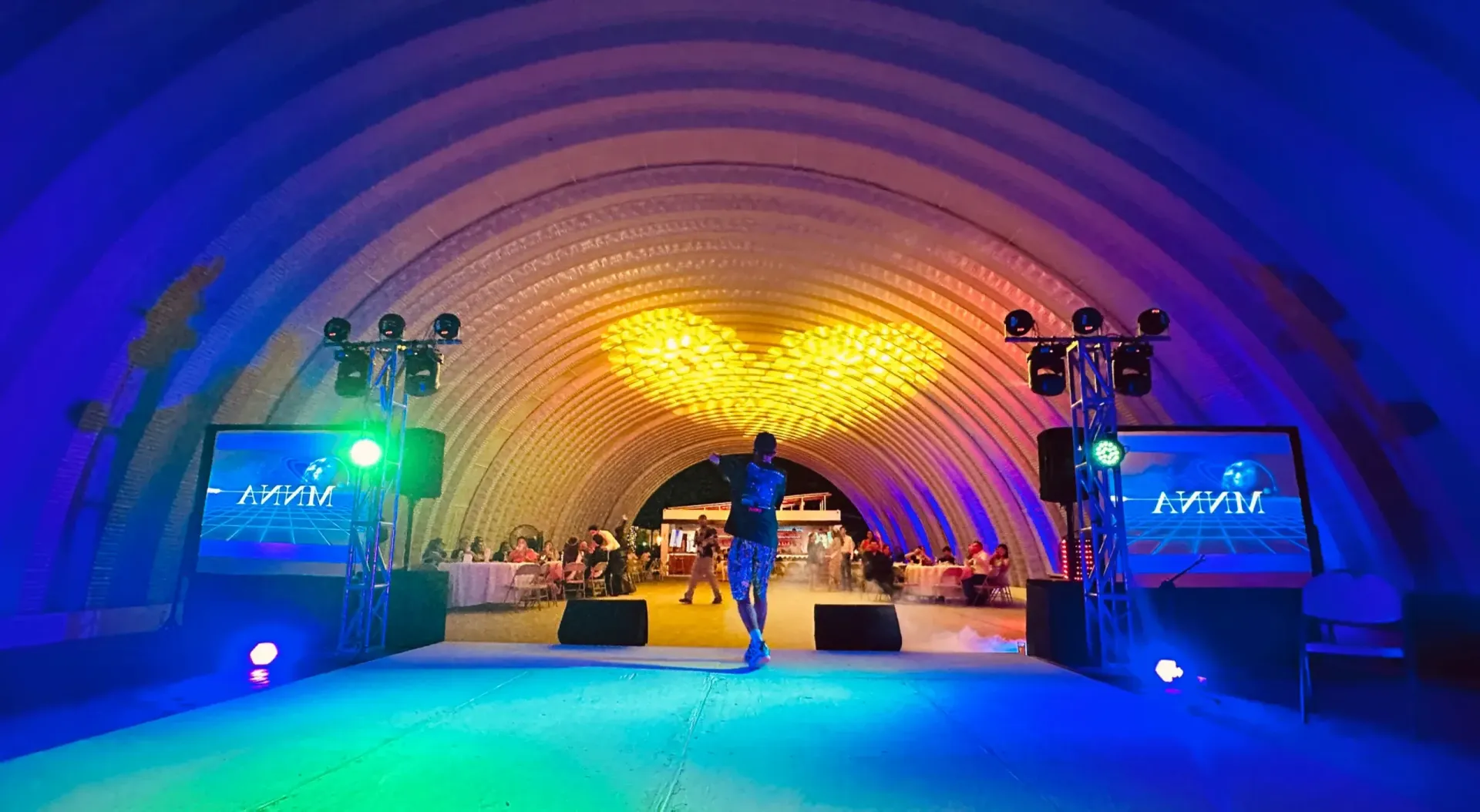 Hombre en el escenario con pantallas, en un colorido recinto con forma de arco. Iluminado con luces azules y naranjas.