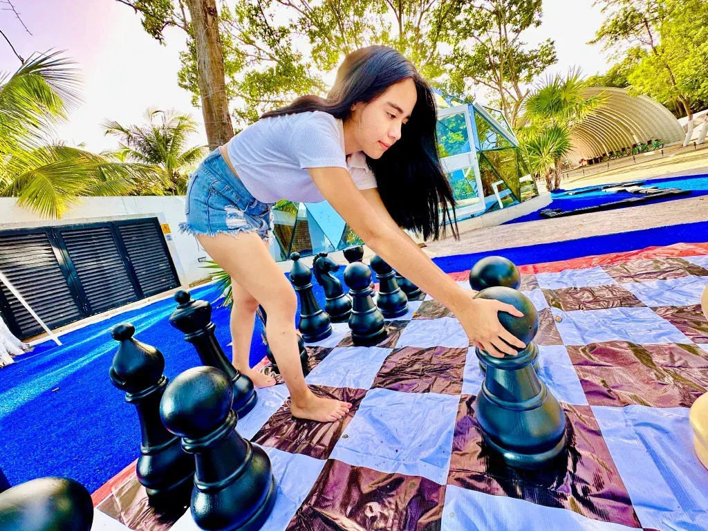 Mujer jugando al ajedrez gigante al aire libre, intentando alcanzar una pieza. Fondo azul y blanco.