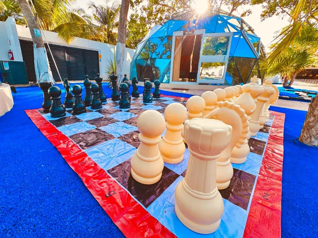 Juego de ajedrez gigante sobre una superficie azul con piezas negras y color crema; estructura de cúpula al fondo.
