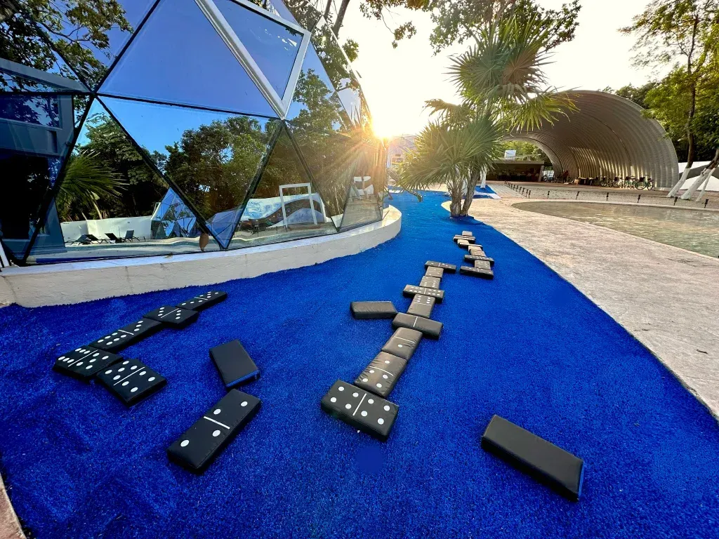 Zona exterior azul con fichas de dominó esparcidas. Edificio moderno con cúpula y cielo soleado al fondo.