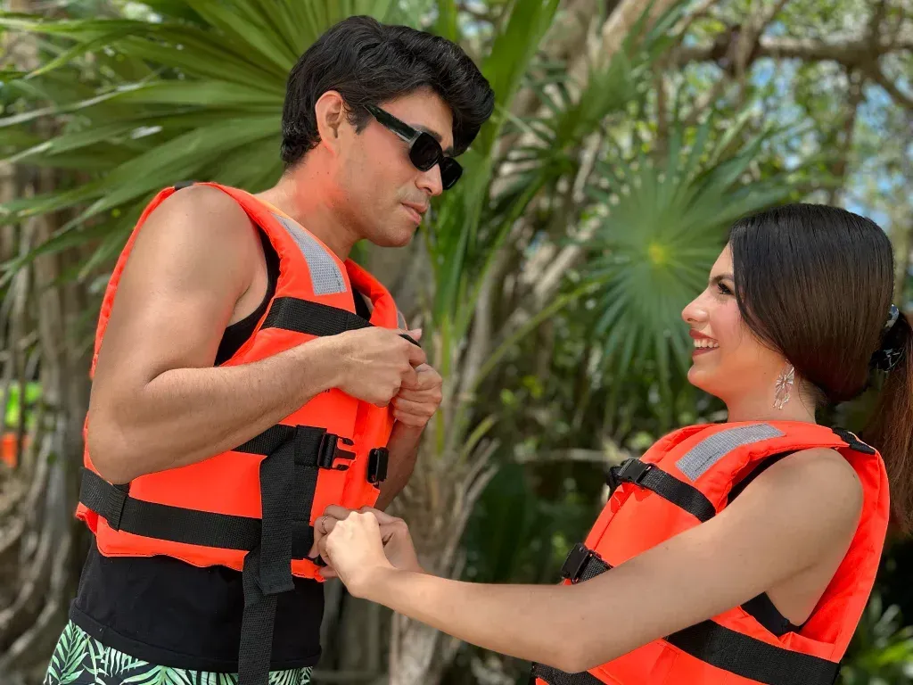 Hombre y mujer con chalecos salvavidas naranjas, sonriendo, al aire libre cerca de vegetación tropical.