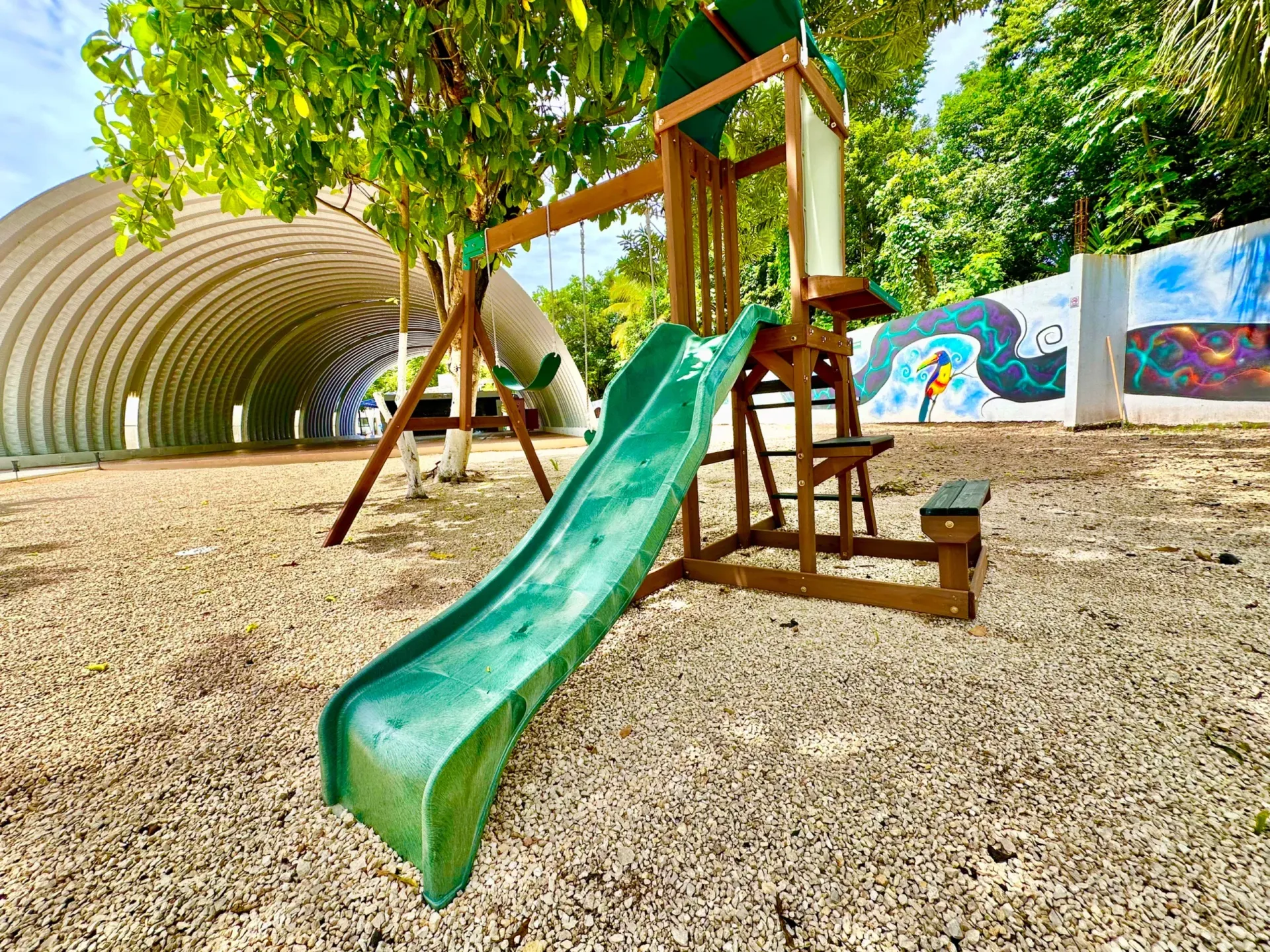 Zona de juegos con tobogán verde y columpios sobre grava, con túnel al fondo.