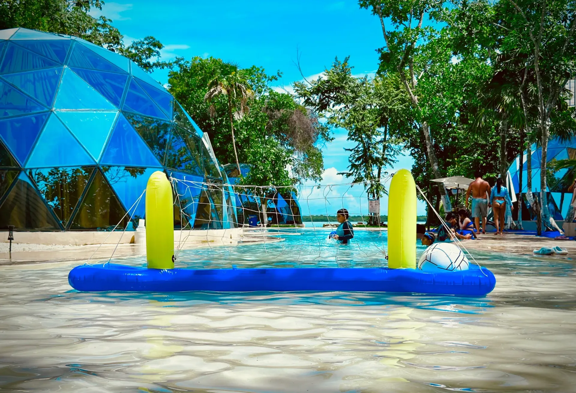 Parque acuático con red de voleibol inflable; gente jugando cerca de una cúpula geodésica azul.