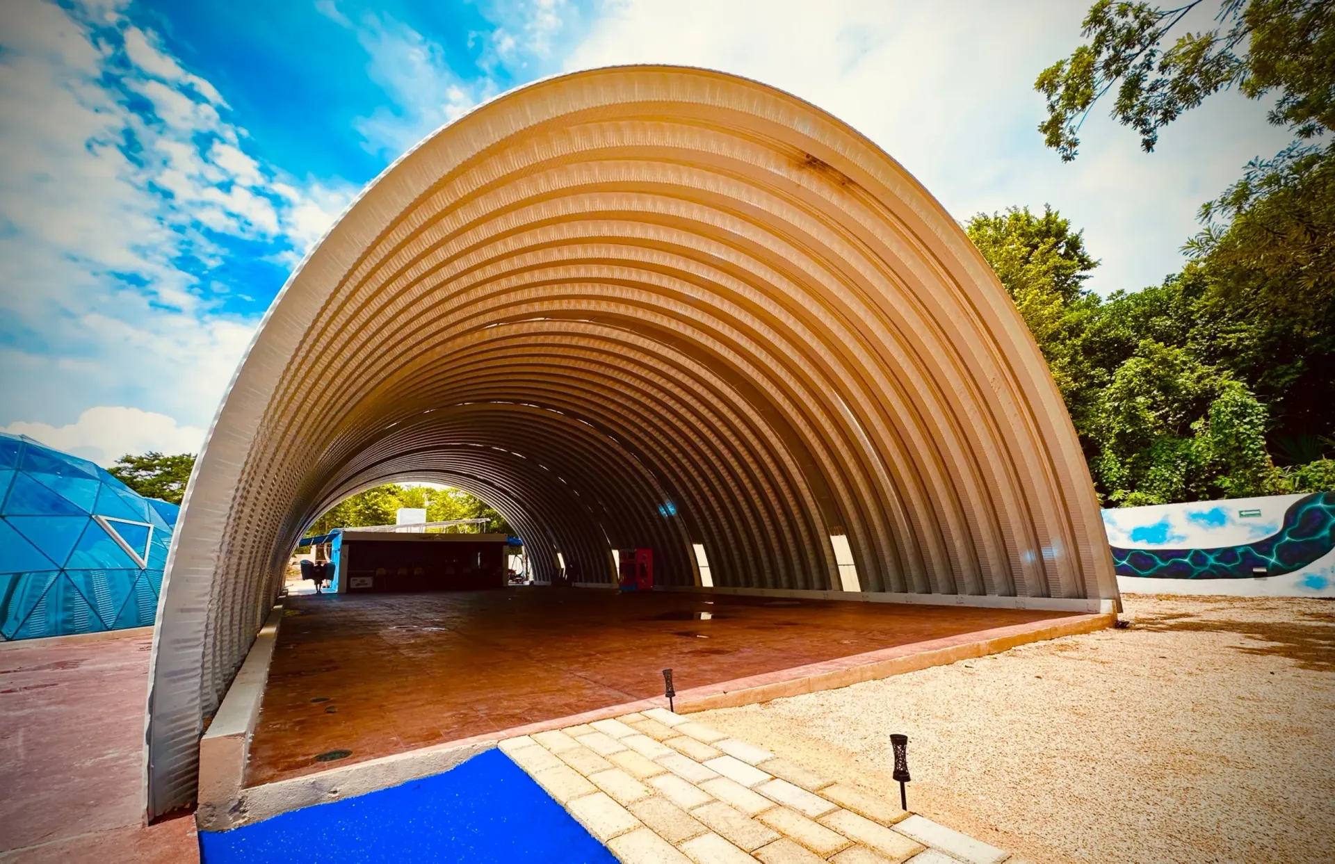 Edificio en forma de arco, con paredes metálicas, suelo marrón, rodeado de árboles y cielo azul.