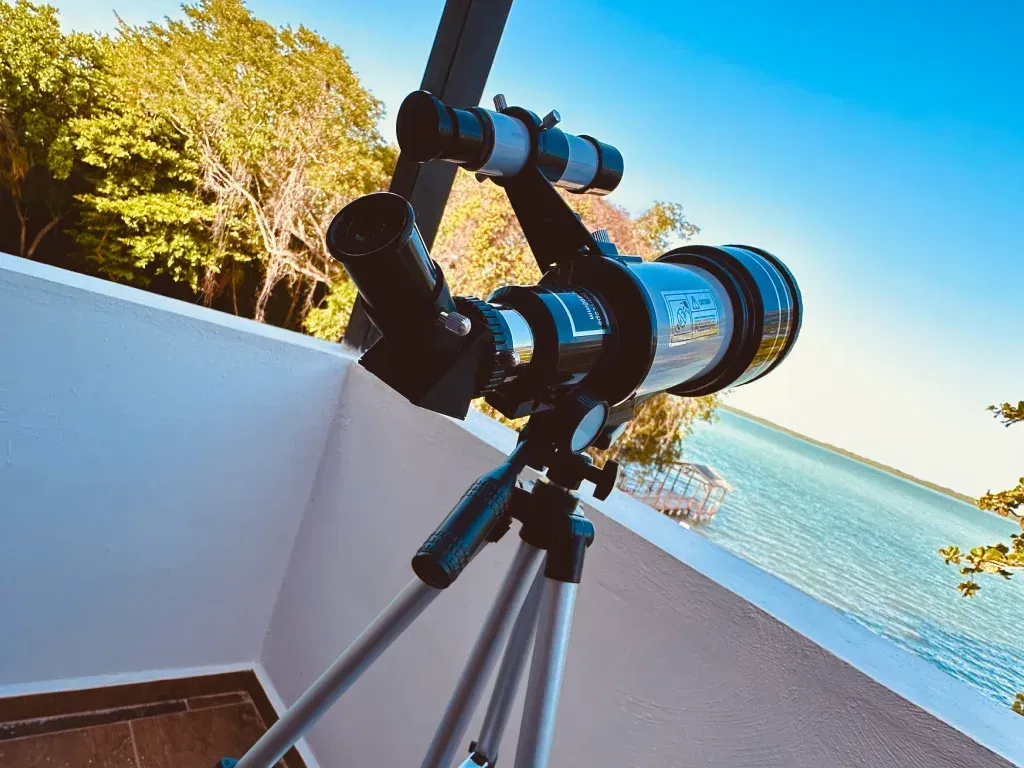 Telescopio sobre trípode, apuntando hacia una masa de agua azul. Luz solar brillante.
