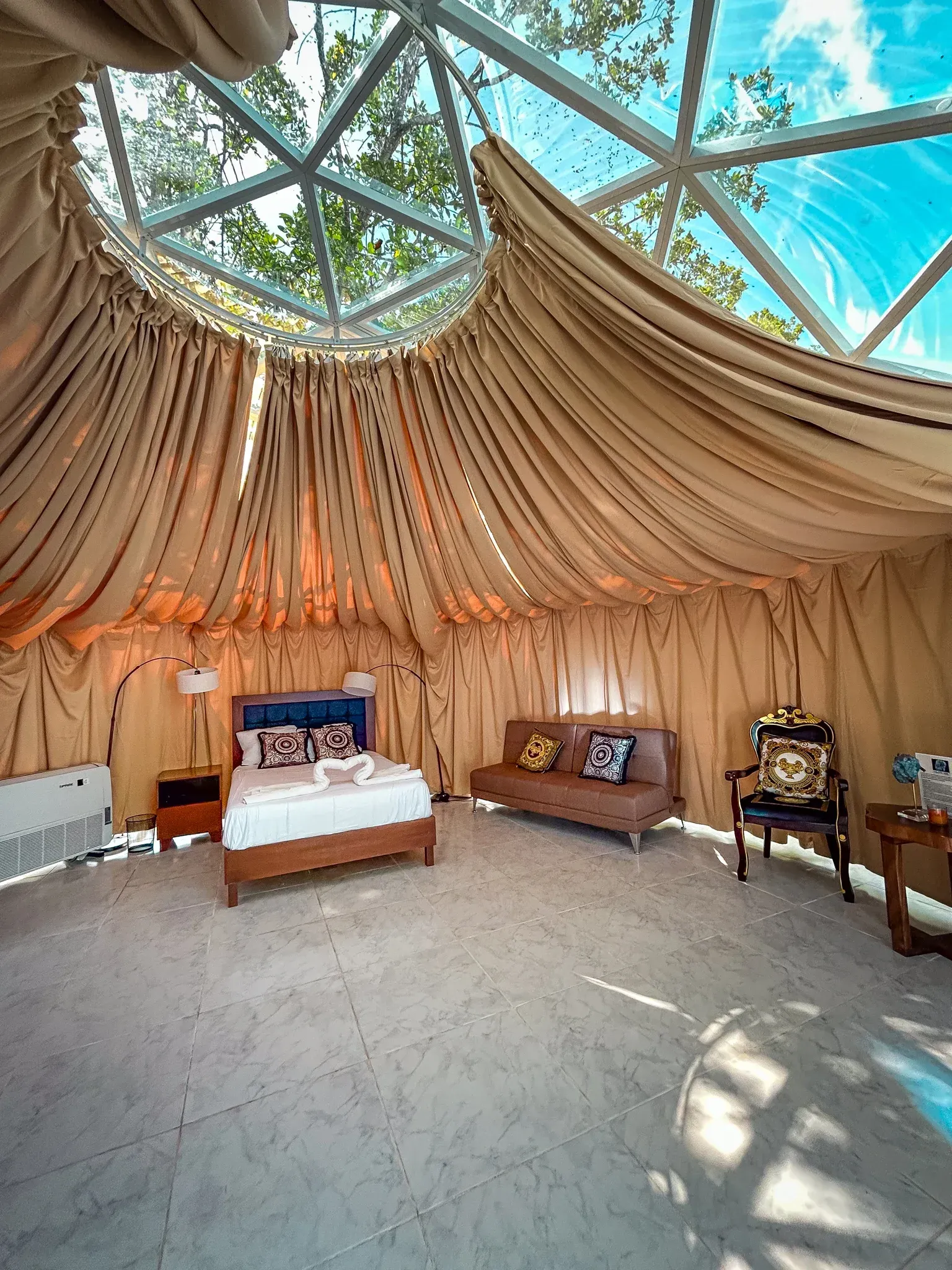 Interior de una habitación en forma de cúpula con una cama, un sofá y una silla bajo un techo transparente.