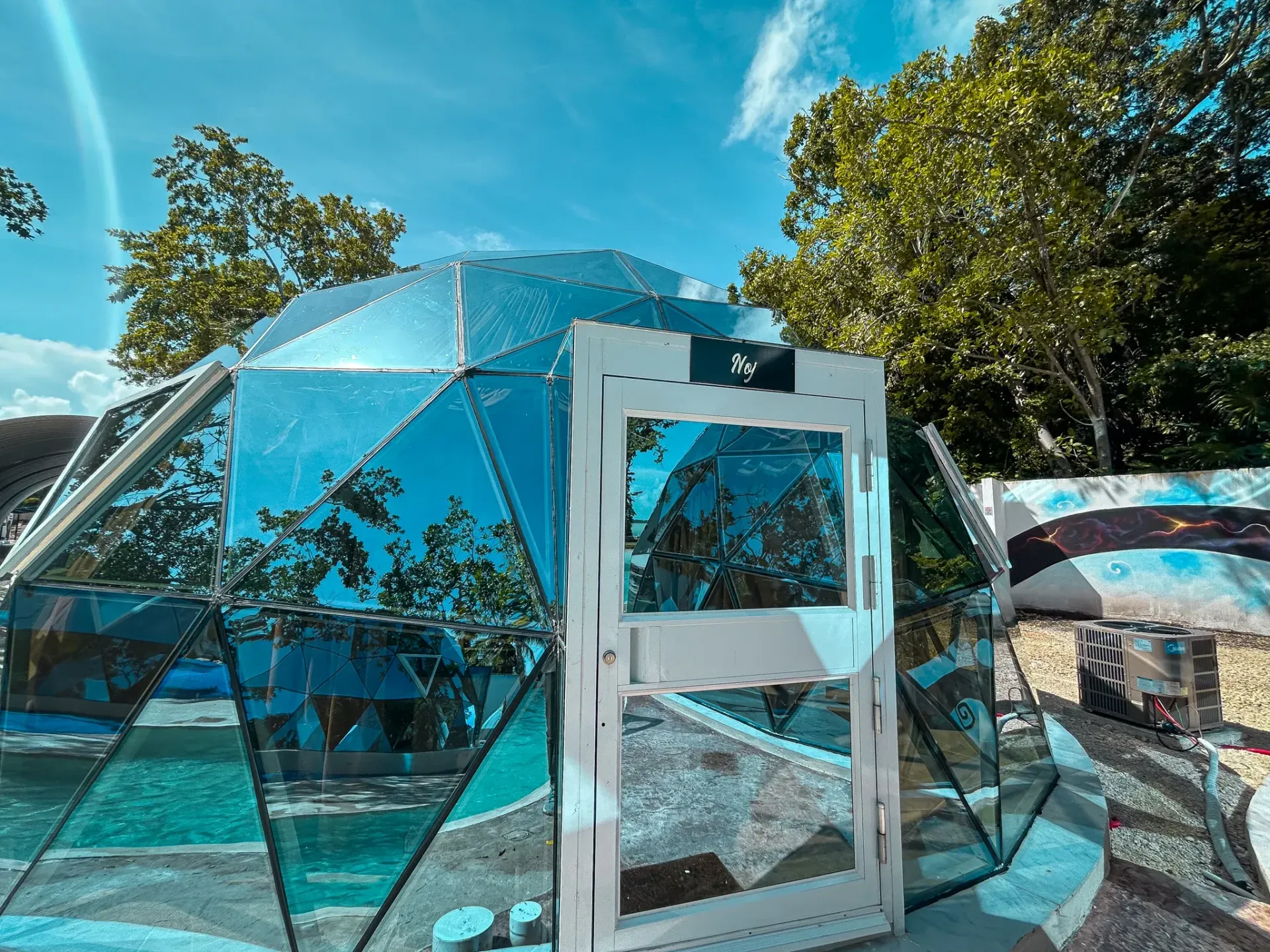 Una estructura espejada en forma de cúpula con una puerta blanca, situada al aire libre bajo un cielo azul.
