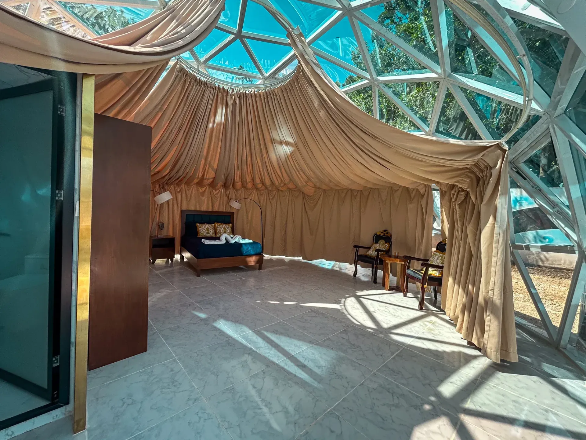 Interior de dormitorio con cúpula geodésica, techo y cama con cortinas. La luz del sol entra a raudales.