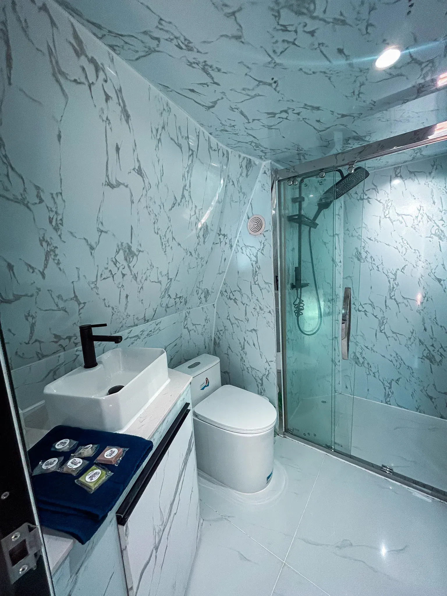 Un baño moderno con paredes y suelo con estampado de mármol. Dispone de lavabo, inodoro y ducha de cristal.