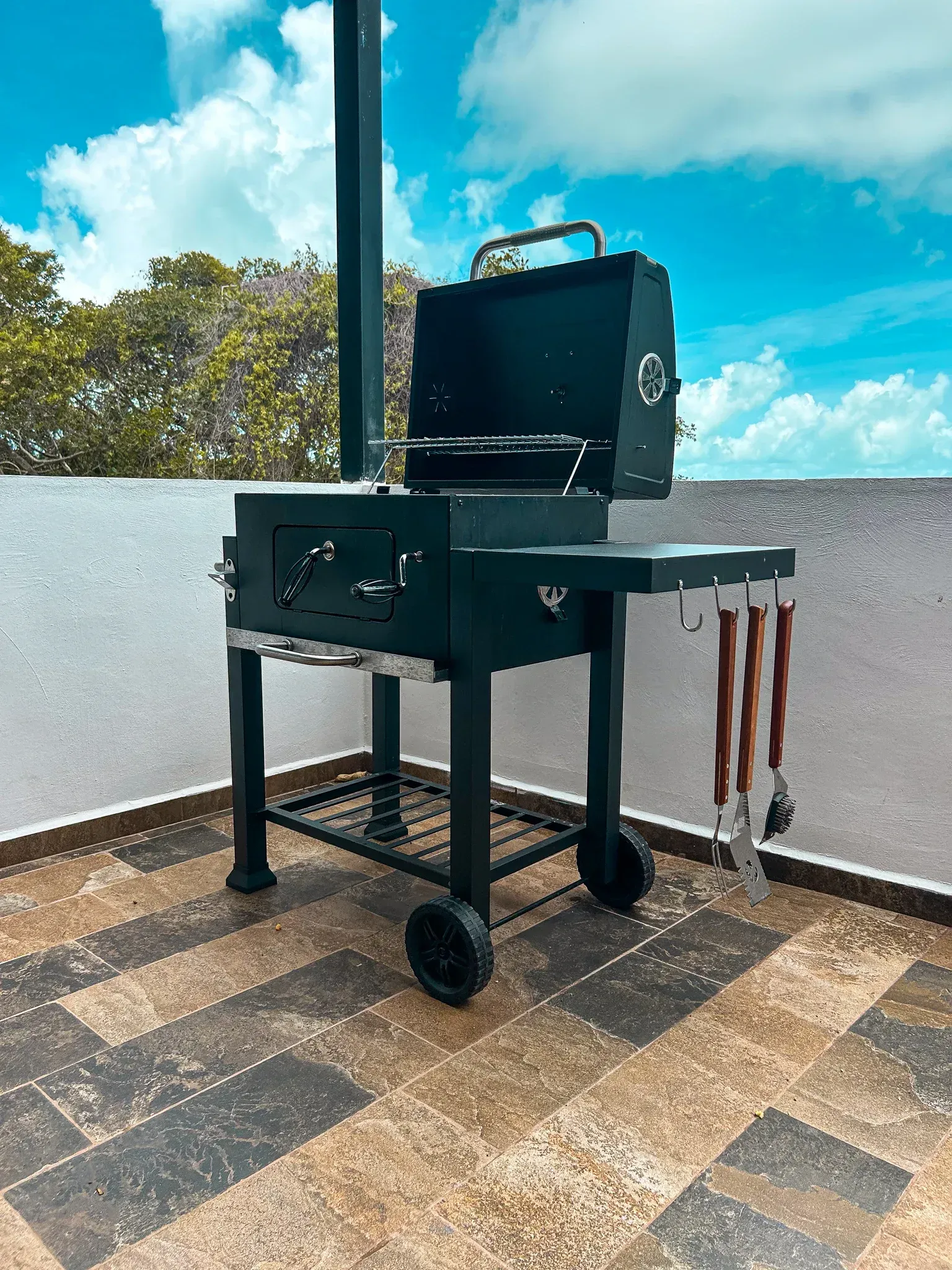 Parrilla de carbón en un patio. De metal negro, con la tapa abierta. Incluye utensilios de cocina colgados.