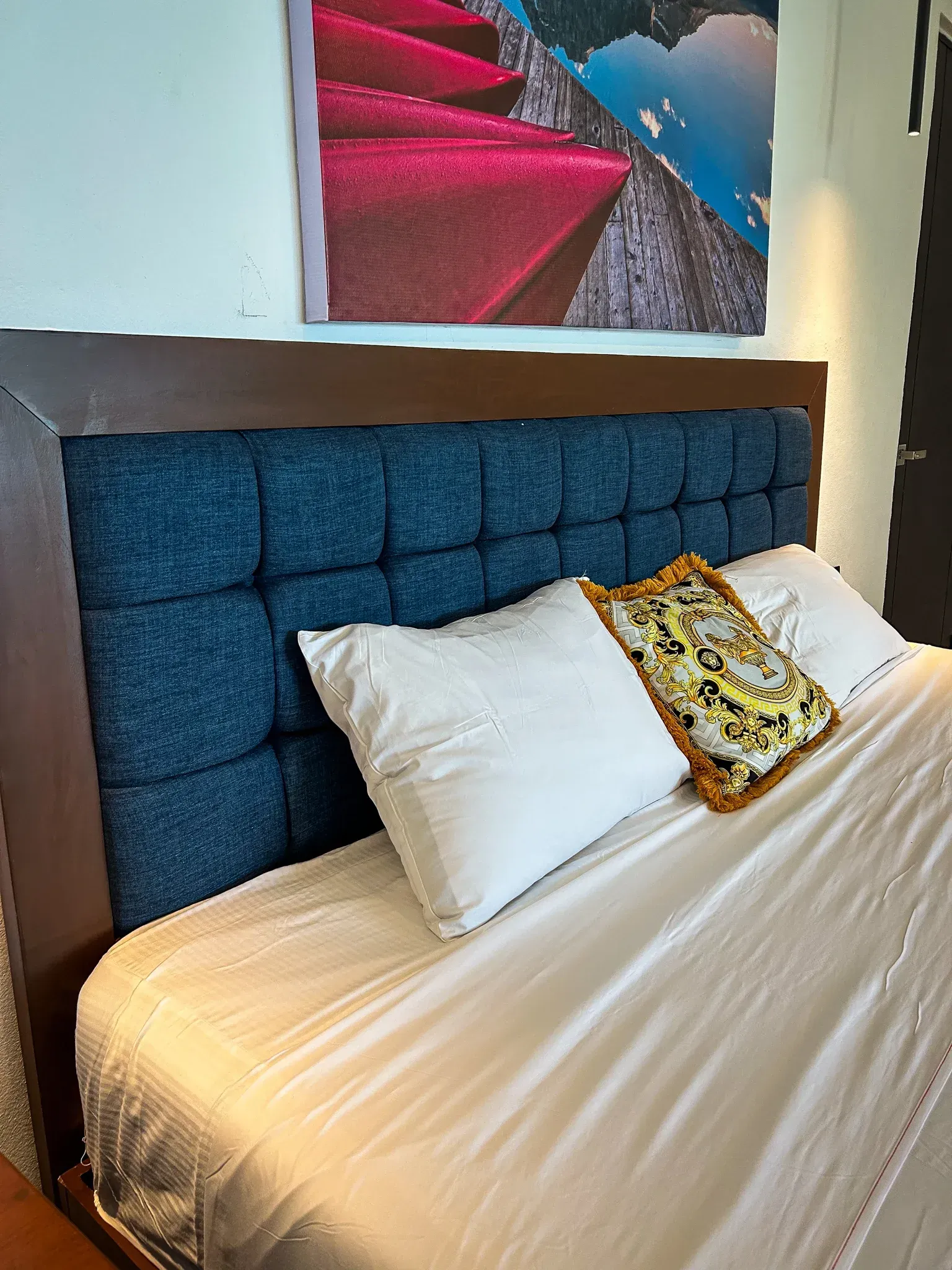 Una cama con cabecero tapizado en azul, sábanas blancas y almohadas. Encima hay un cuadro en la pared.