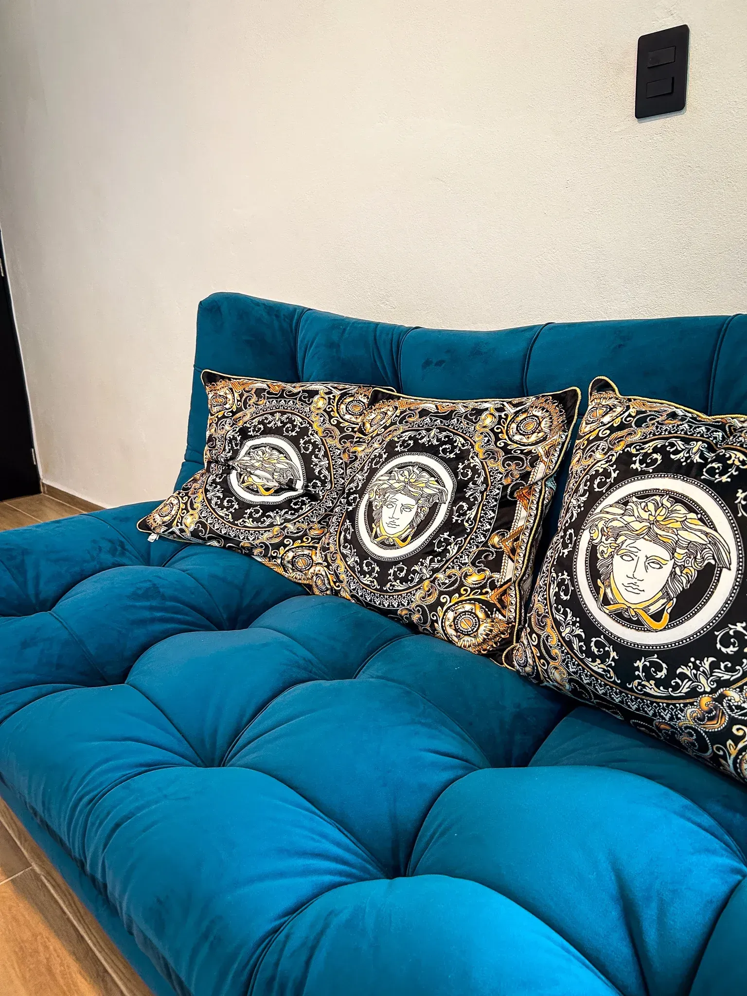 Sofá capitoné color verde azulado con dos cojines Versace. Pared blanca, enchufe negro.