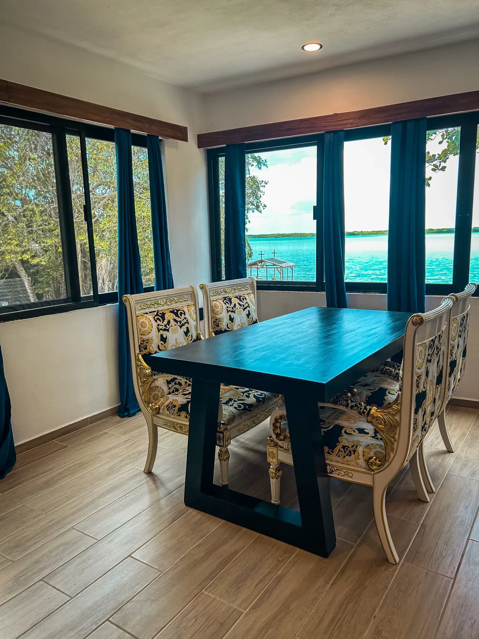 Comedor con mesa negra, sillas estampadas y grandes ventanales con vistas al agua.