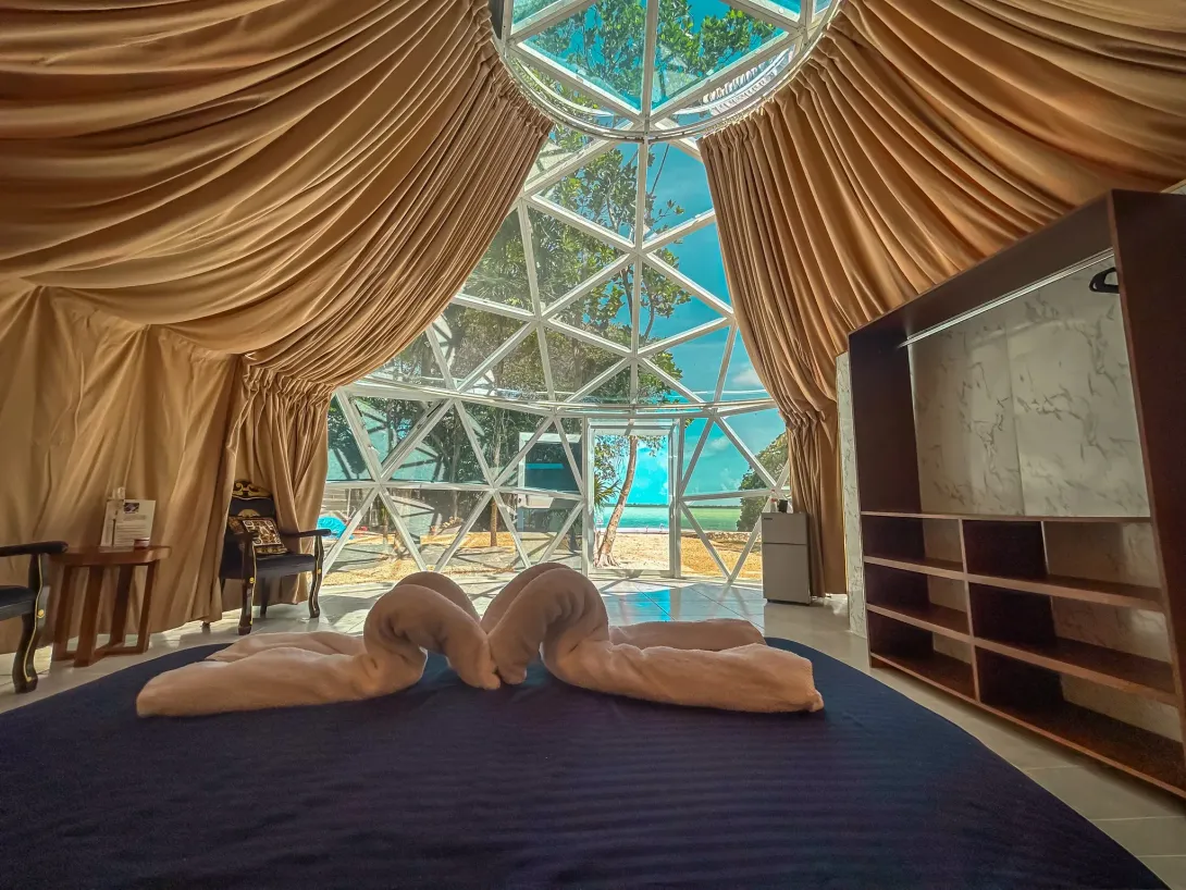 Interior de una lujosa tienda de campaña en forma de cúpula con toallas de cisne sobre una cama y vistas al océano.