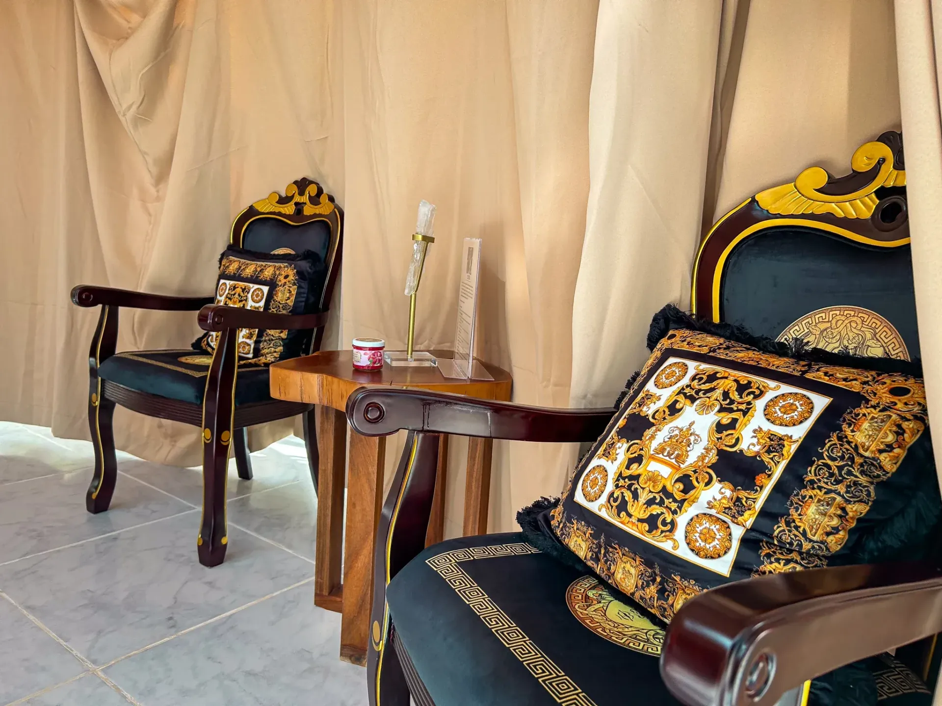 Dos sillas ornamentadas con cojines decorativos, una mesita de madera, cortinas vaporosas, suelo de baldosas.