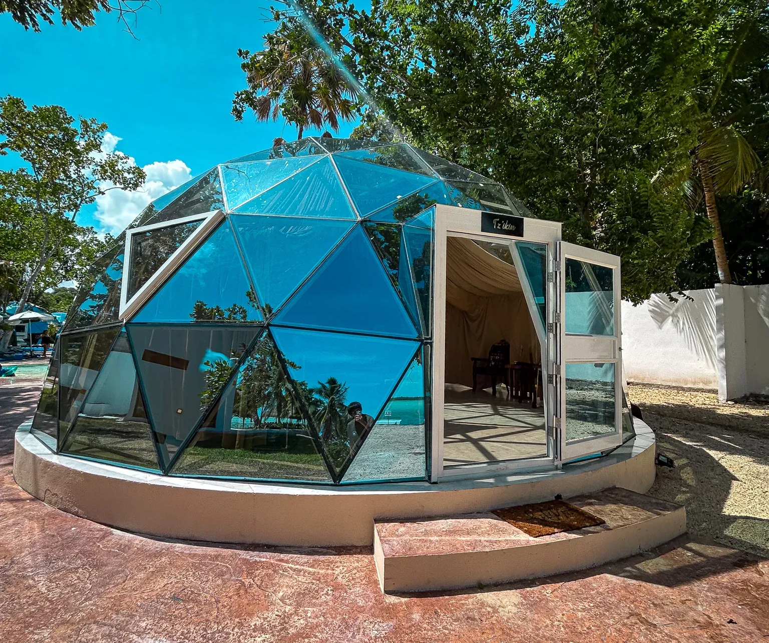 Edificio con cúpula geodésica, paneles de vidrio reflectante y puertas abiertas, ubicado en un espacio exterior soleado.