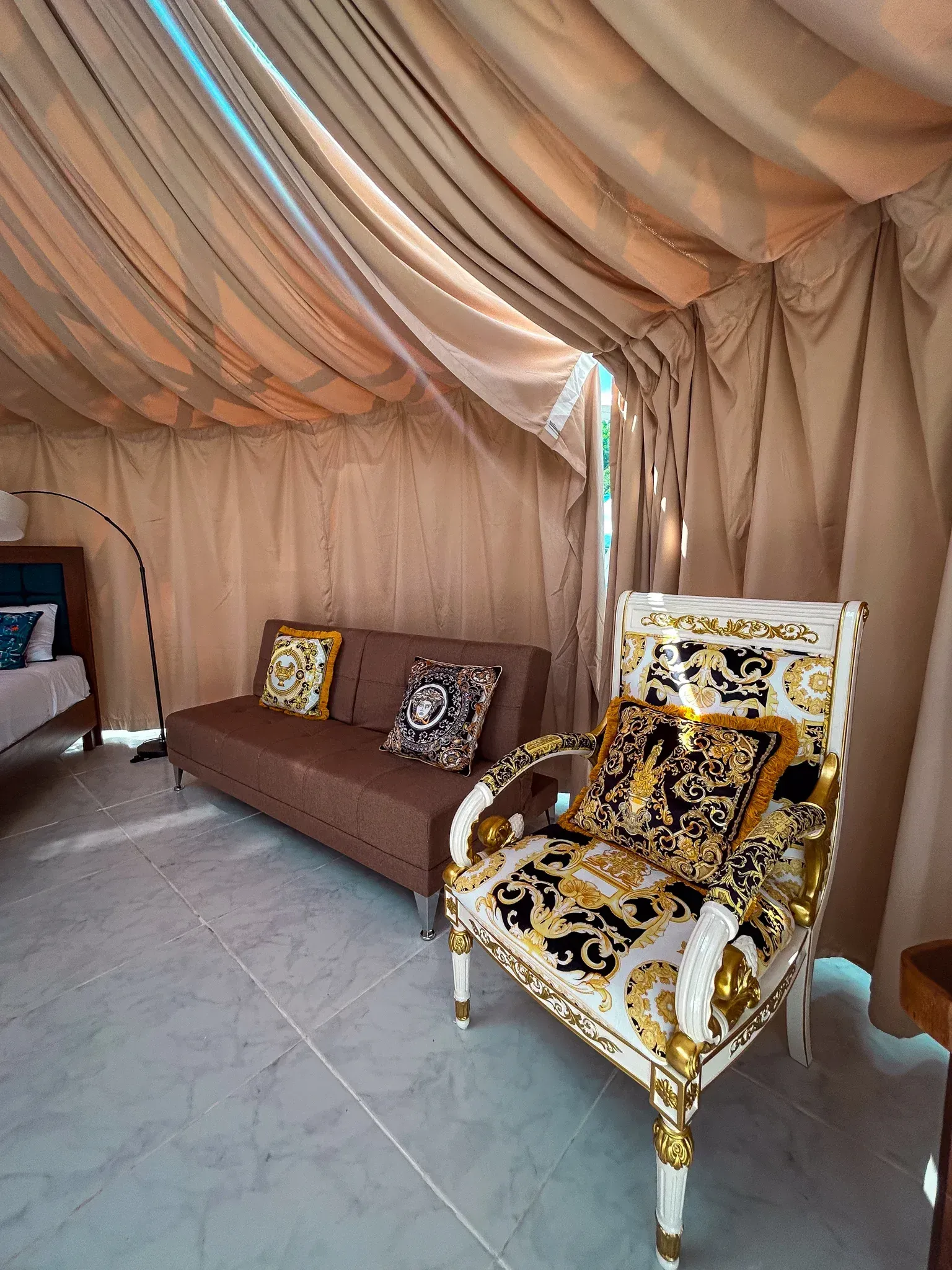 Interior de una lujosa tienda de campaña con sofá marrón, silla blanca ornamentada, cortinas beige y un atisbo de una cama.