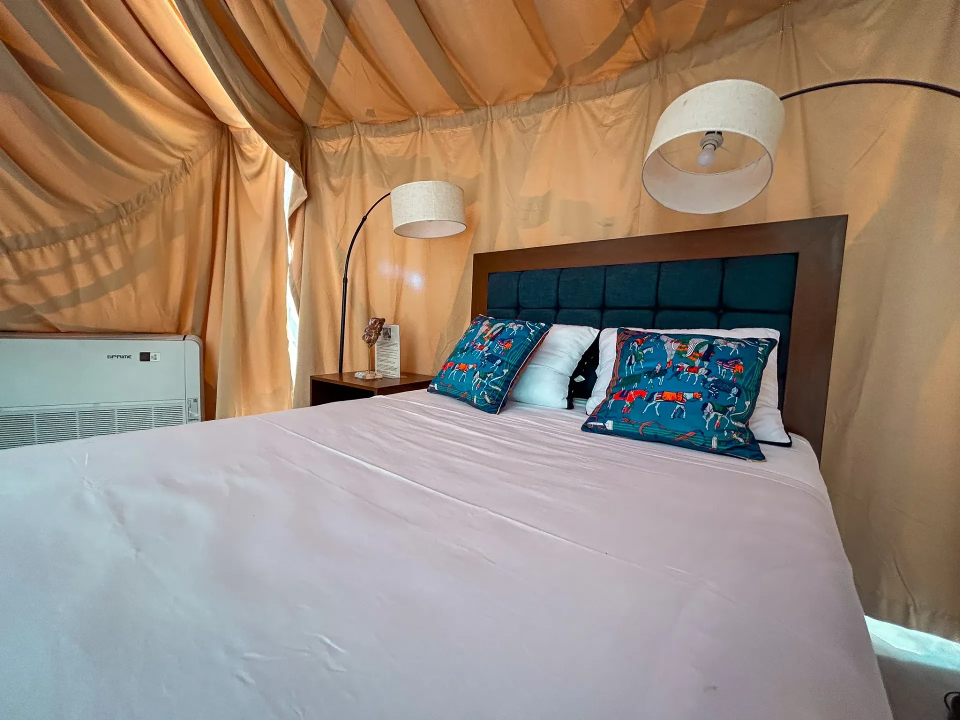 Interior de dormitorio acogedor con cama, almohadas y lámparas. Paredes color beige y cabecero de madera.