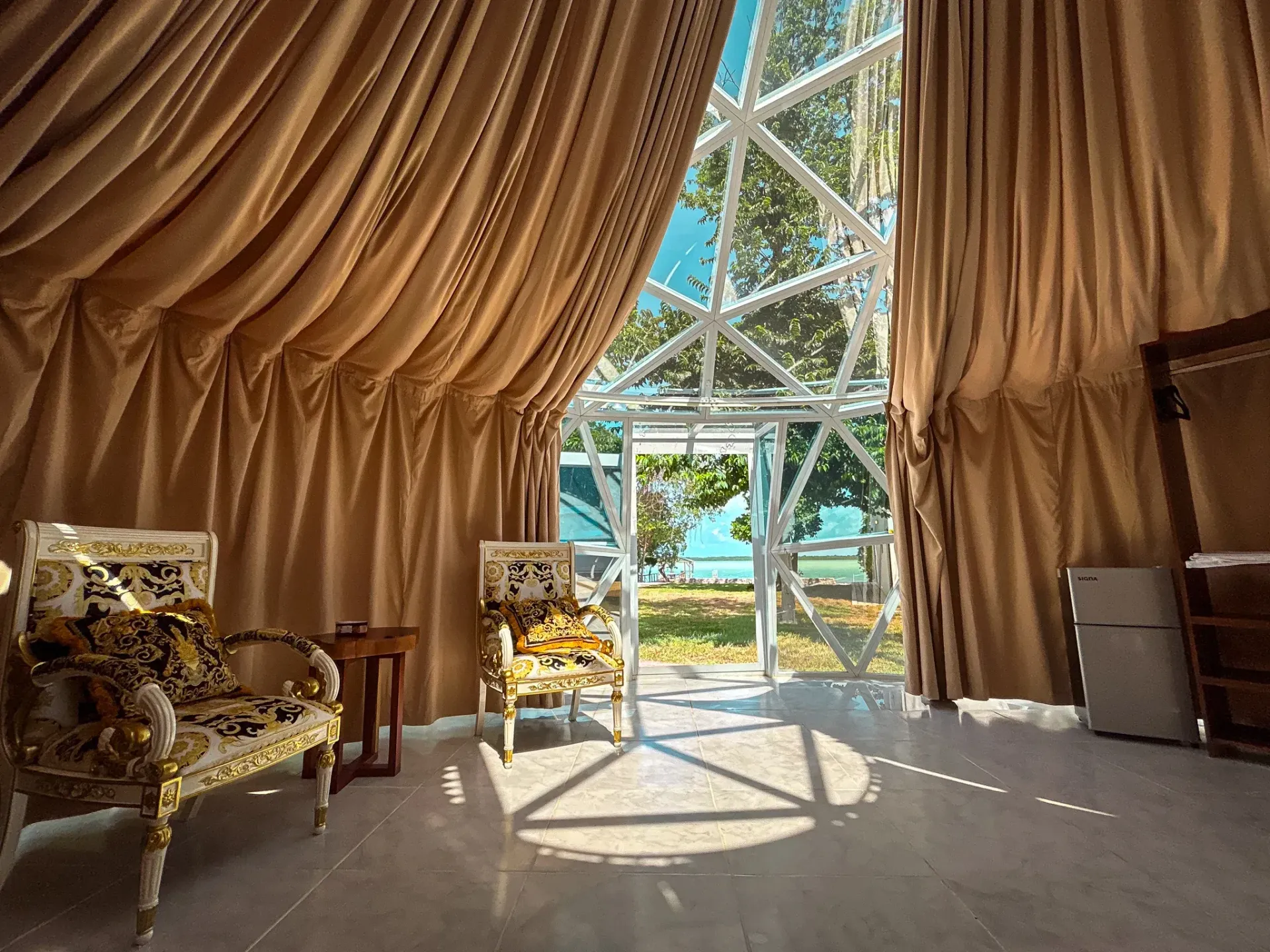 Interior con cortinas color canela, dos sillas ornamentadas y una puerta de cristal geométrica que da a una vista al agua.