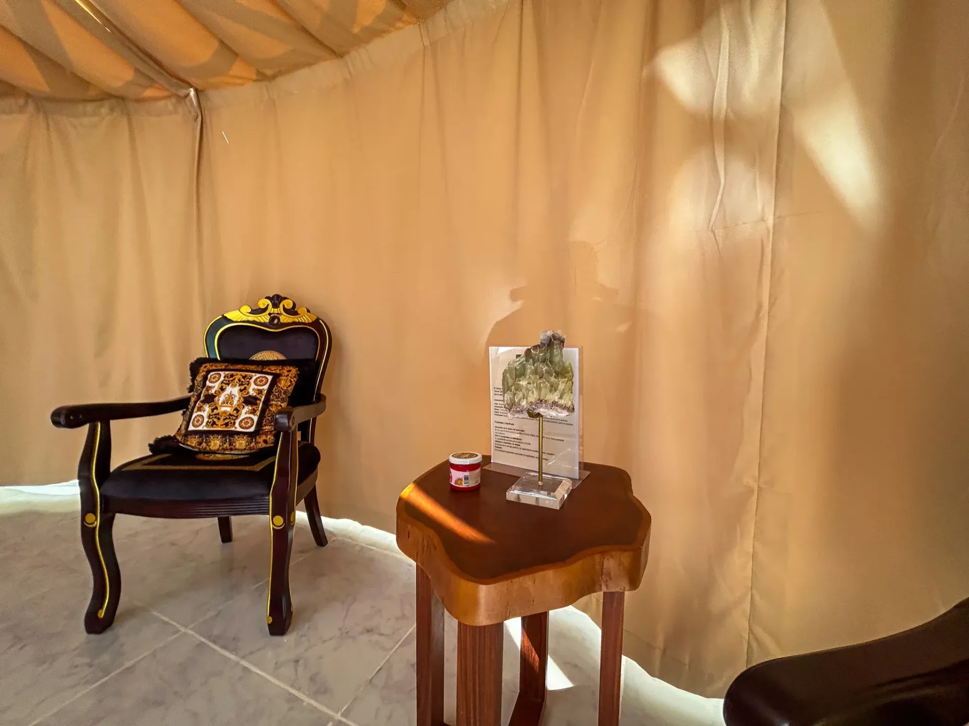 Dentro de una estructura parecida a una tienda de campaña, una silla oscura y ornamentada y una pequeña mesa de madera.