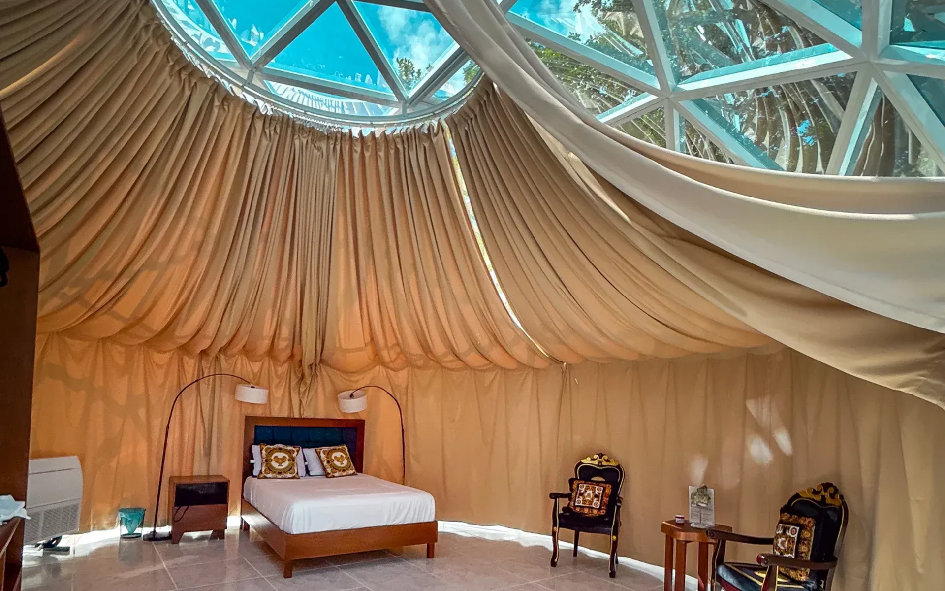 Dormitorio con paredes curvas de color beige, una cama, sillas estampadas y un techo abovedado de cristal.