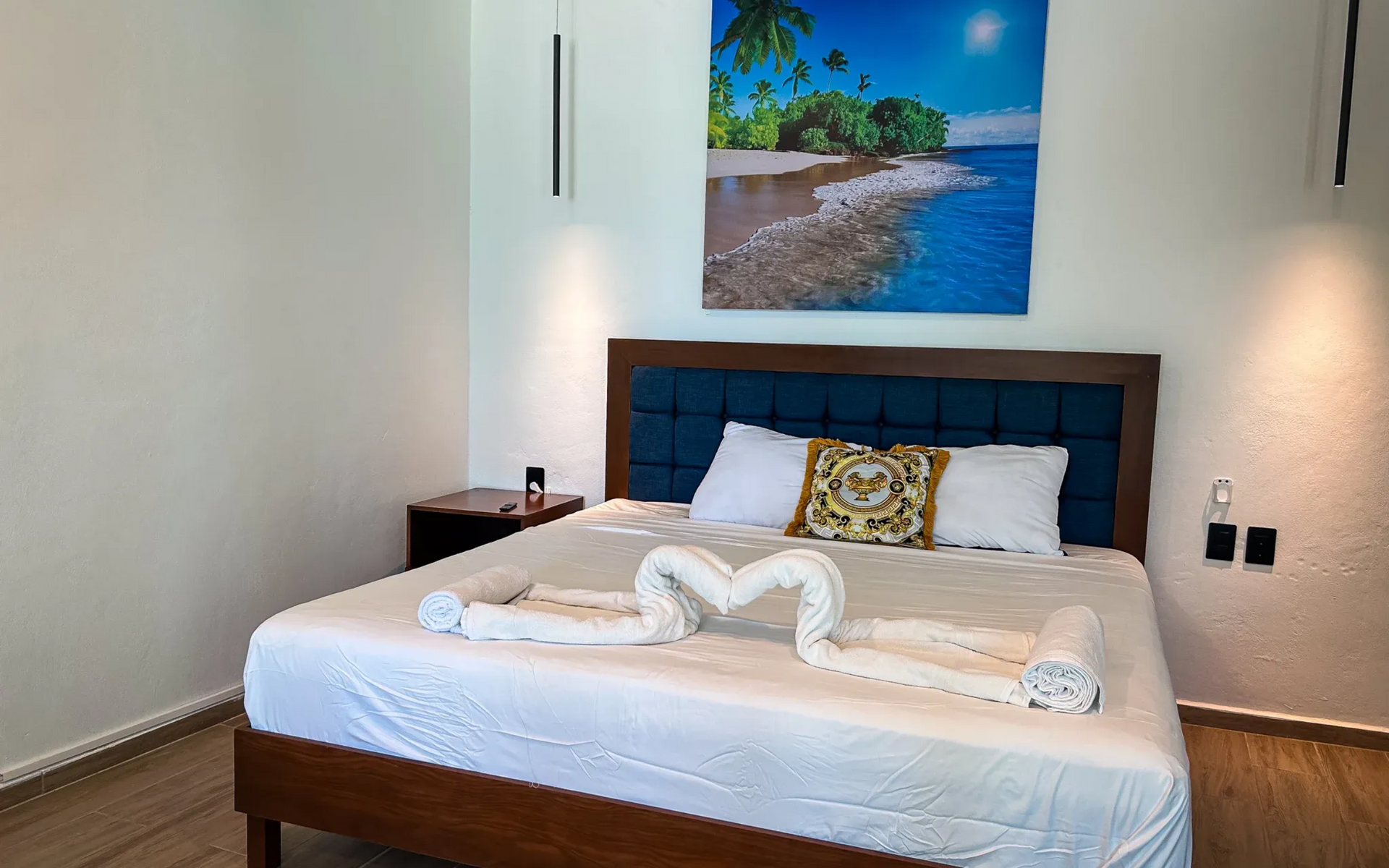 Dormitorio con cama, cabecero azul, sábanas blancas y un cuadro de una playa en la pared.